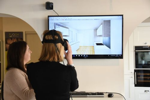 a customer tryin gout the virtual reality headset with her kitchen design whilst the designer stands next to her watching on the screen 